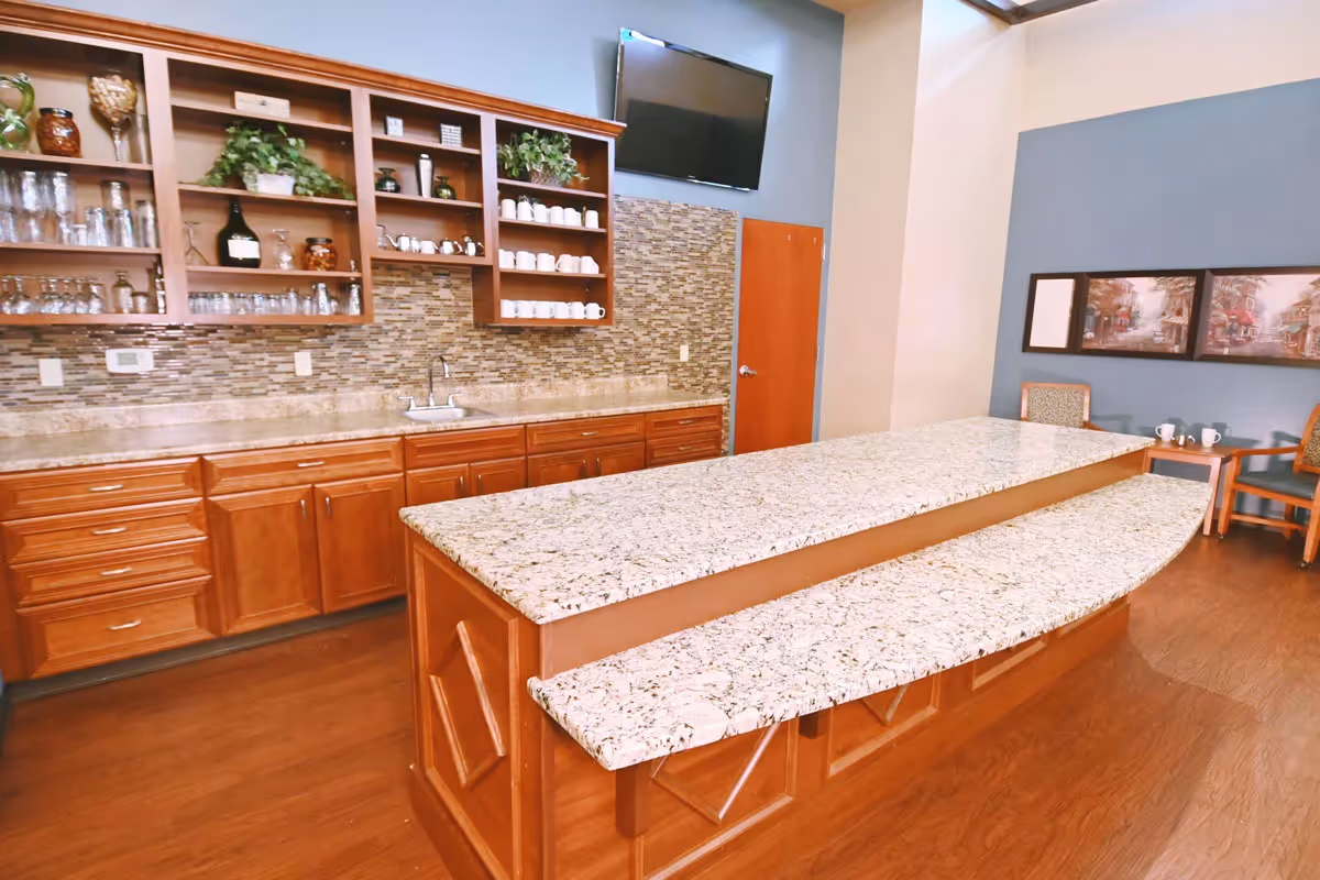 Communal kitchen area with a long granite island, wooden cabinets and open shelving holding glassware and mugs, and a wall-mounted TV.