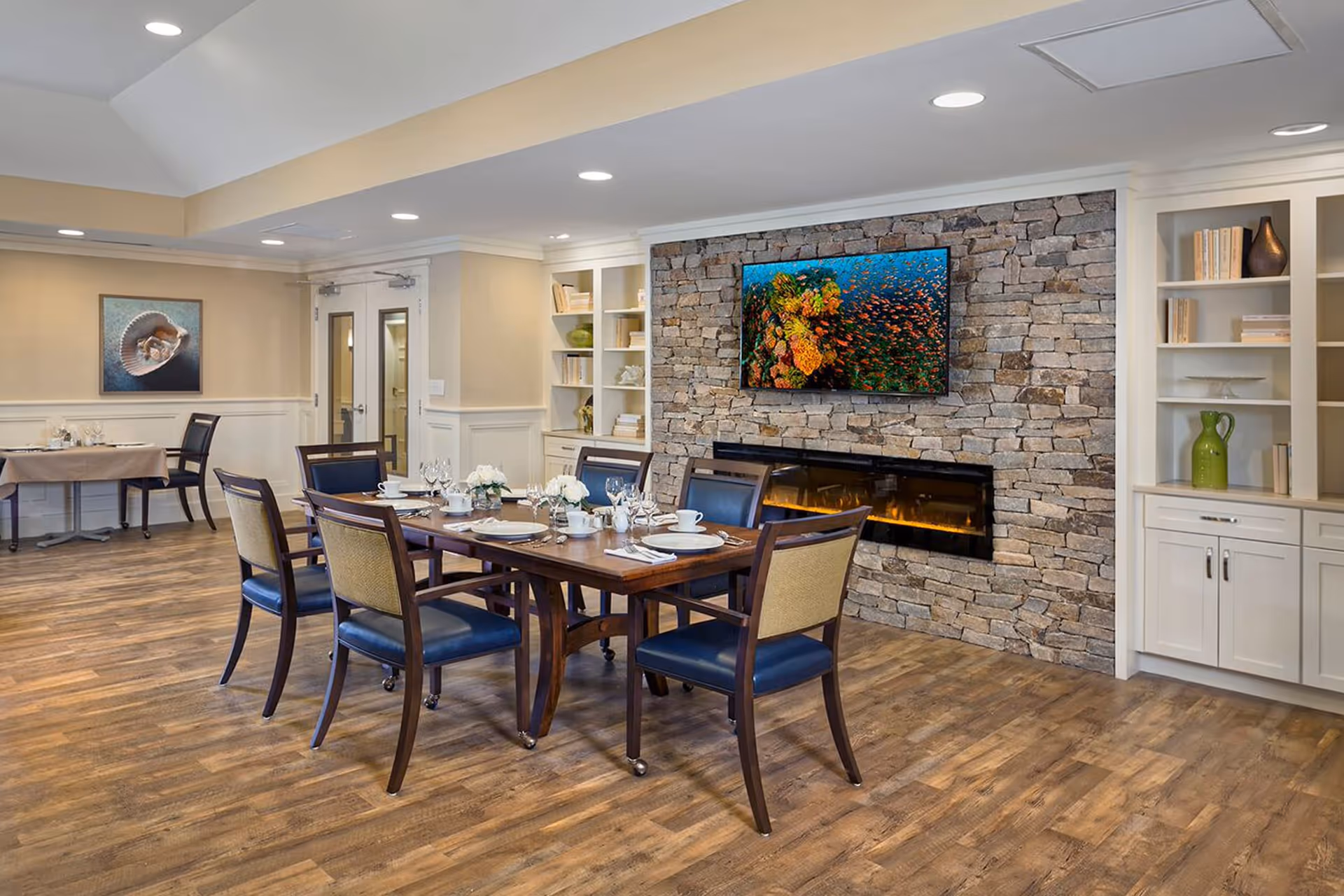 A dining room in a senior living facility with a wooden table set for six people with plates, cups, and napkins. The room features a stone accent wall with a mounted flat-screen TV displaying an underwater coral reef scene above a modern electric fireplace. There are built-in white shelves with decorative items and books on either side of the fireplace. The floor is wood, and the walls are painted beige with white trim. Additional tables and chairs are visible in the background.