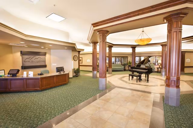 Spacious and well-lit interior lobby area of Rocky Mountain Care - Hunter Hollow featuring a reception desk on the left, a grand piano in the center under a decorative ceiling with wooden columns, green carpeted flooring, and seating areas in the background.