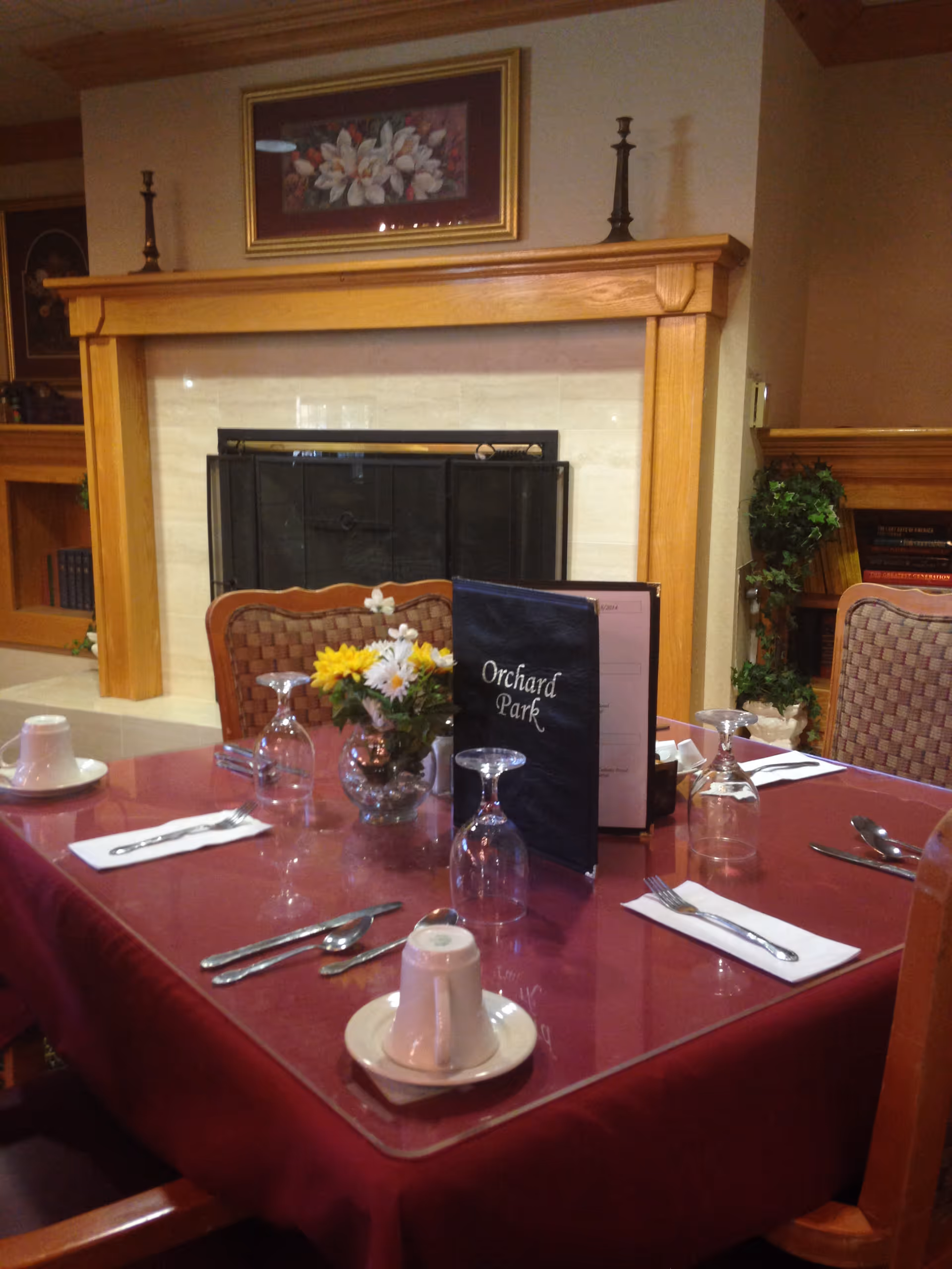 A dining table set for four with a red tablecloth, white cups and saucers, silverware, and upside-down wine glasses. A small vase with yellow and white flowers and a menu labeled 'Orchard Park' are on the table. In the background, there is a fireplace with a wooden mantel, two candlesticks, and a framed floral painting above it. There are also bookshelves and a potted plant visible.