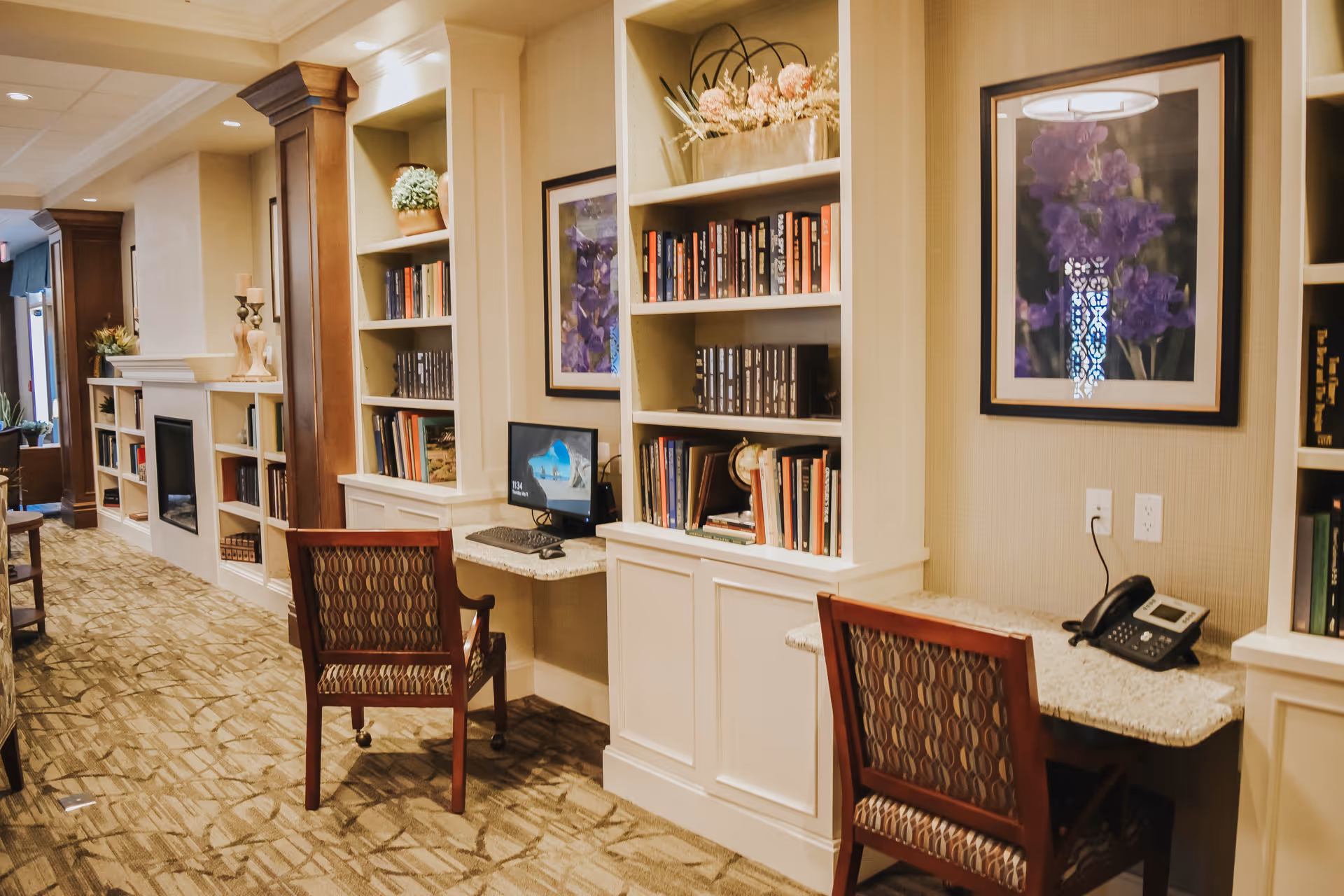 Interior view of a senior living facility hallway with built-in bookshelves, two desks with chairs, a computer, a telephone, and framed floral artwork on the walls. The space is warmly lit with carpeted flooring and decorative wooden columns.