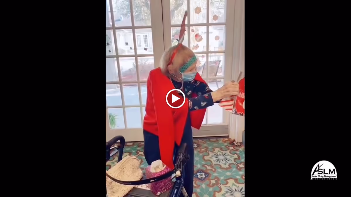 An elderly woman wearing a red cape, a festive headband with reindeer antlers, and a face mask is standing indoors near a glass door decorated with holiday stickers. She is holding a red and white striped gift bag and using a walker. The floor has a colorful patterned carpet.