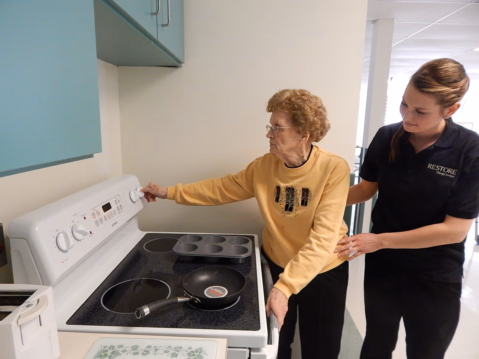 An elderly woman wearing a yellow sweater is adjusting the knob on a white electric stove in a kitchen while a younger woman in a black Restore Therapy Services shirt supports her from behind.