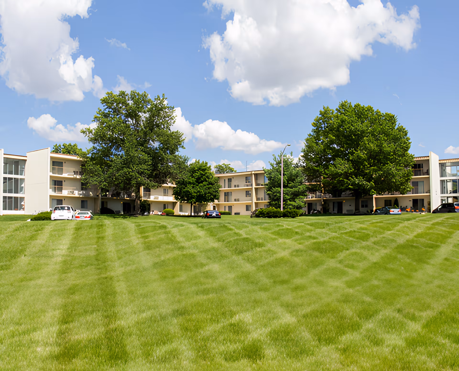 Low-rise senior living buildings with balconies and trees behind a wide manicured lawn under a blue sky.