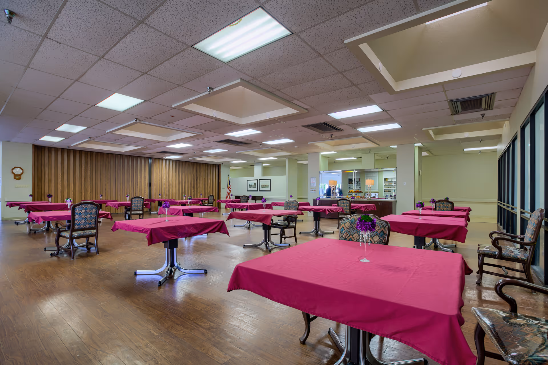A spacious dining room with multiple tables covered in red tablecloths, each adorned with a small vase of purple flowers. The room has wooden floors, patterned chairs, and a ceiling with recessed lighting. There is a wooden partition on one side and large windows on the other.