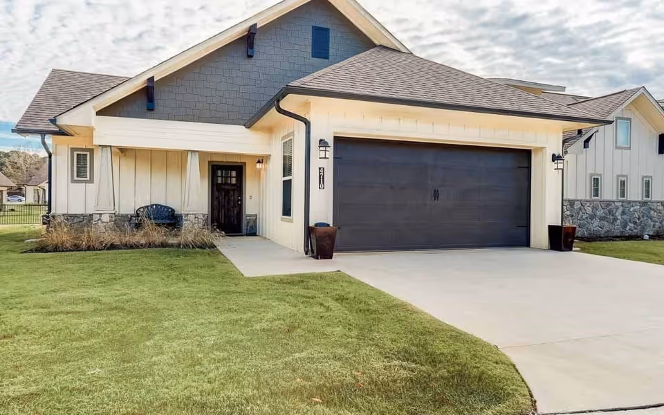 Exterior view of a modern single-story house with a dark gray garage door, light-colored siding, stone accents, and a small covered porch with a bench. The house is surrounded by a well-maintained green lawn and a concrete driveway leading to the garage.