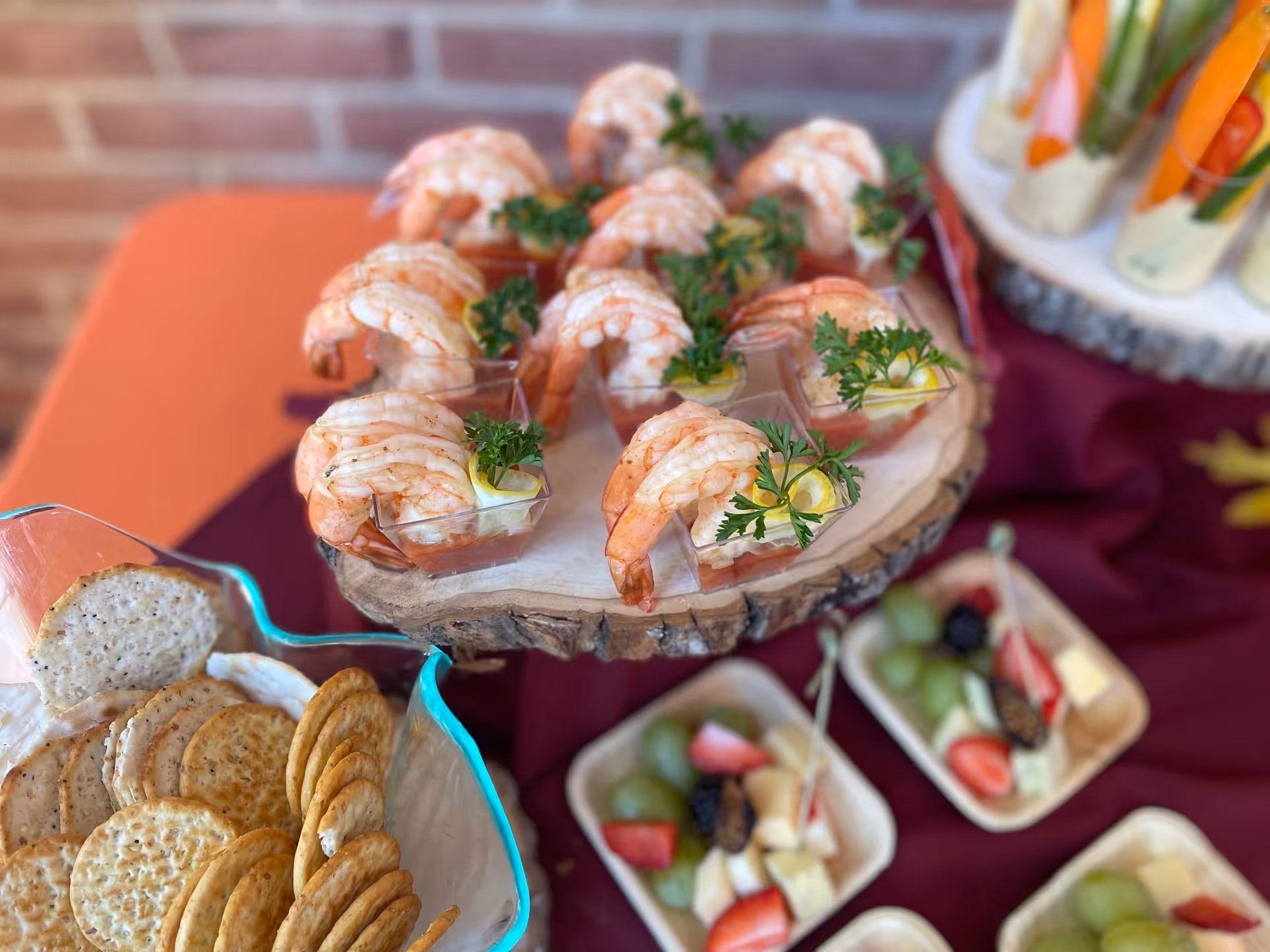 Close-up of a food display featuring shrimp cocktail served in small clear cups with lemon wedges and parsley on a wooden platter. Surrounding the platter are plates with crackers and small servings of fruit and cheese. The background includes a brick wall and a table covered with a burgundy cloth.