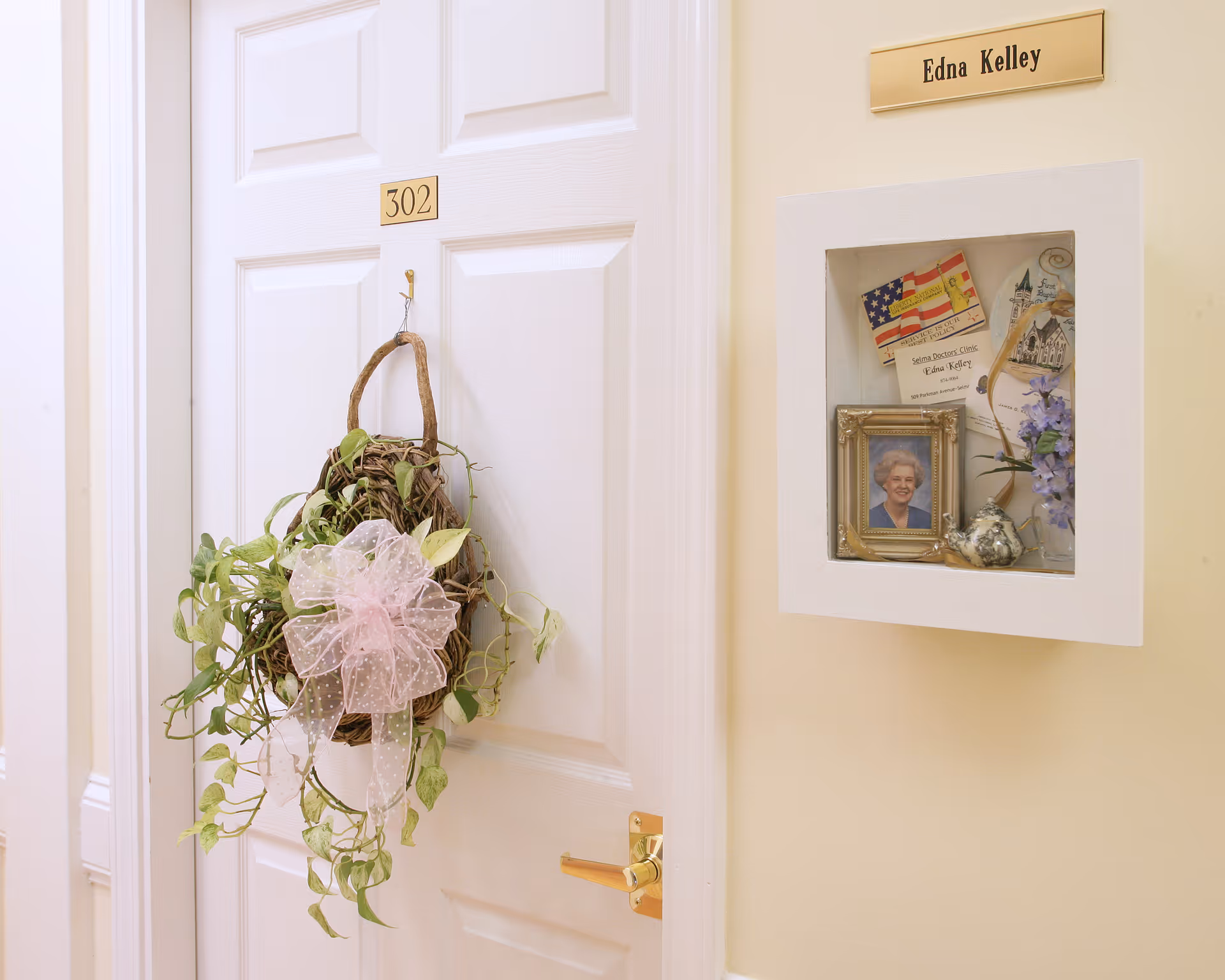 A white door labeled 302 with a decorative hanging wreath made of vines and a pink bow. Next to the door is a small recessed display case on a beige wall containing a framed photo of an elderly woman, a small silver teapot, a vase with purple flowers, and some papers. Above the display case is a nameplate that reads 'Edna Kelley'.