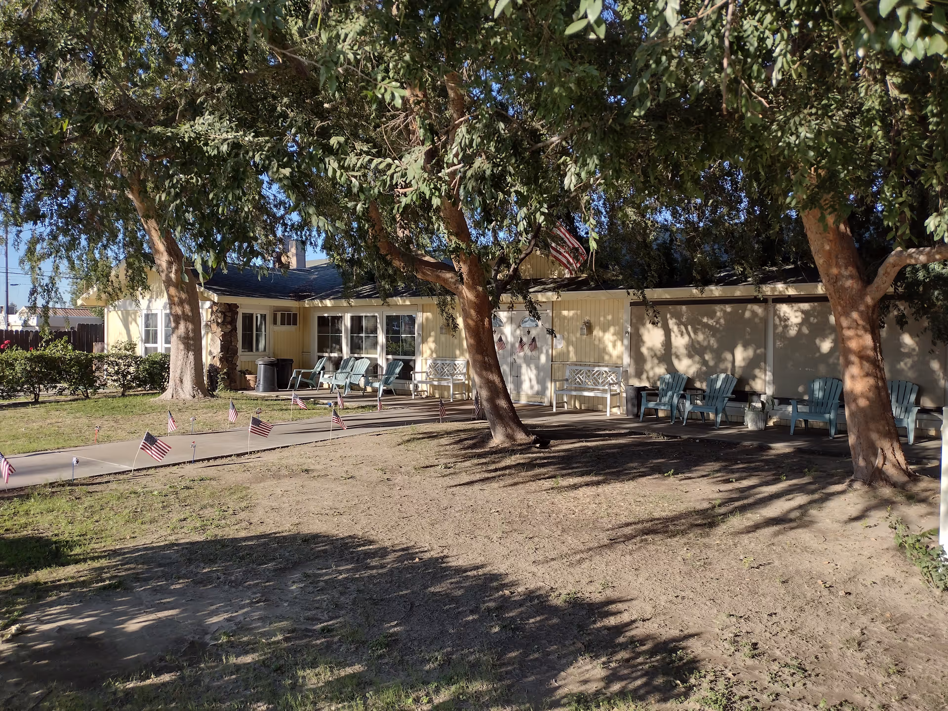 Single-story building front with a shaded lawn, a walkway lined with small American flags, and outdoor chairs under trees.