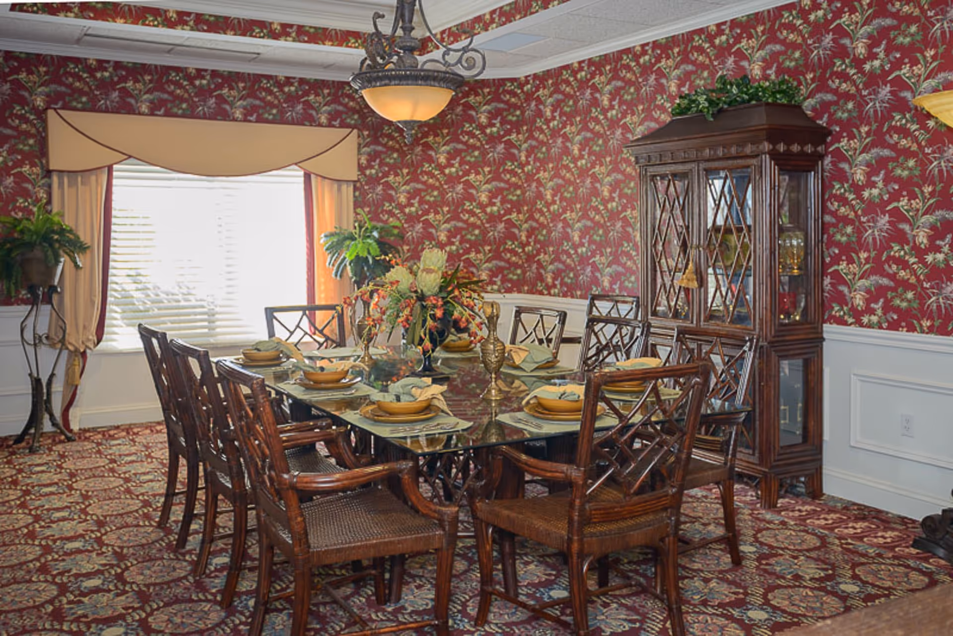 Formal dining room with a glass-top table set for eight, floral wallpaper, china cabinet and patterned carpet.