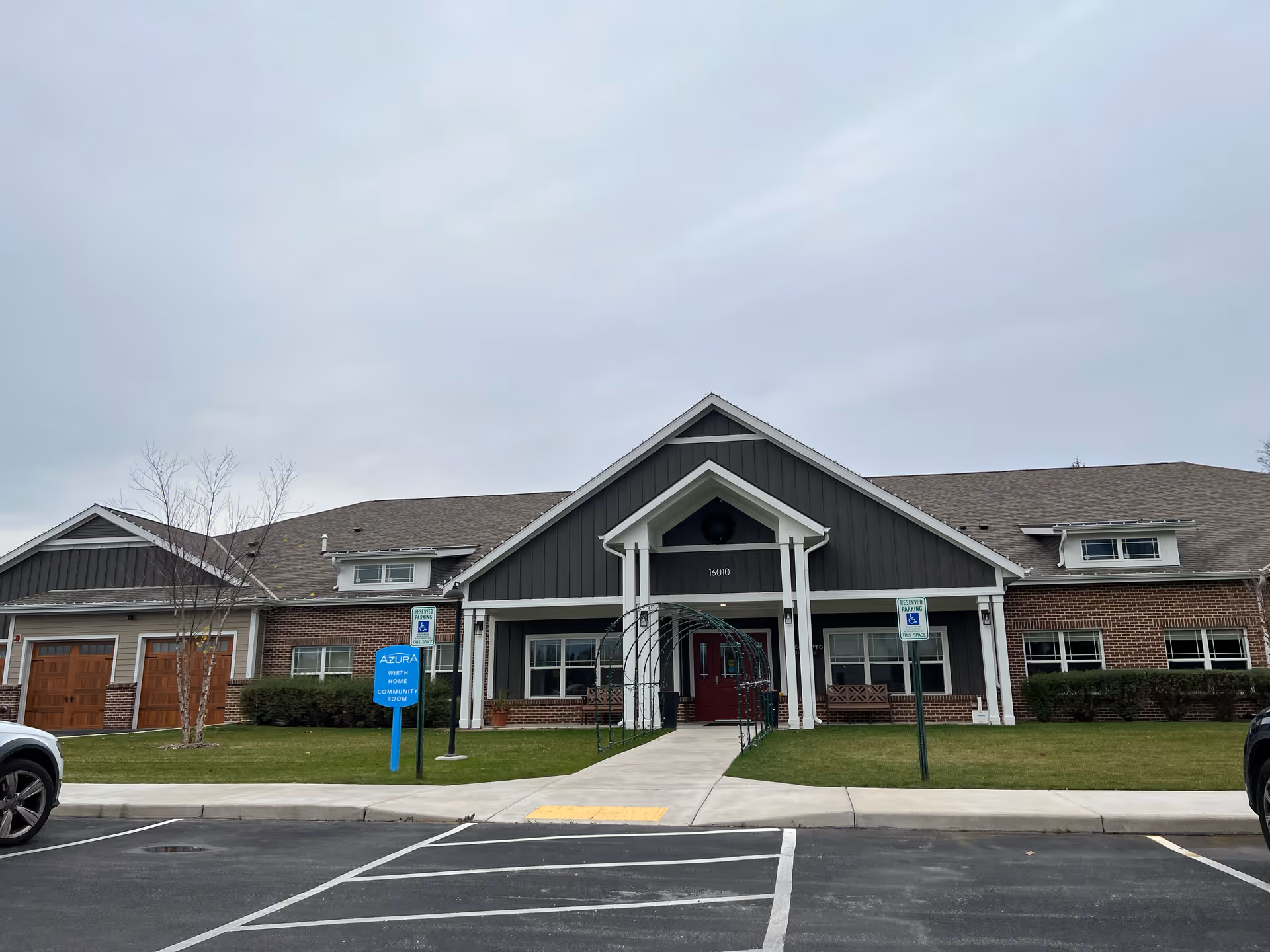 Front exterior view of a single-story building with a peaked roof and a covered entrance supported by white columns. The building has a combination of dark siding and red brick walls, with a red door at the entrance. There is a blue sign on the lawn that reads 'Azura Wirth Home Community Room' and two reserved parking signs for handicapped spaces near the entrance. The sky is overcast and there are a few parked cars visible at the edges of the image.