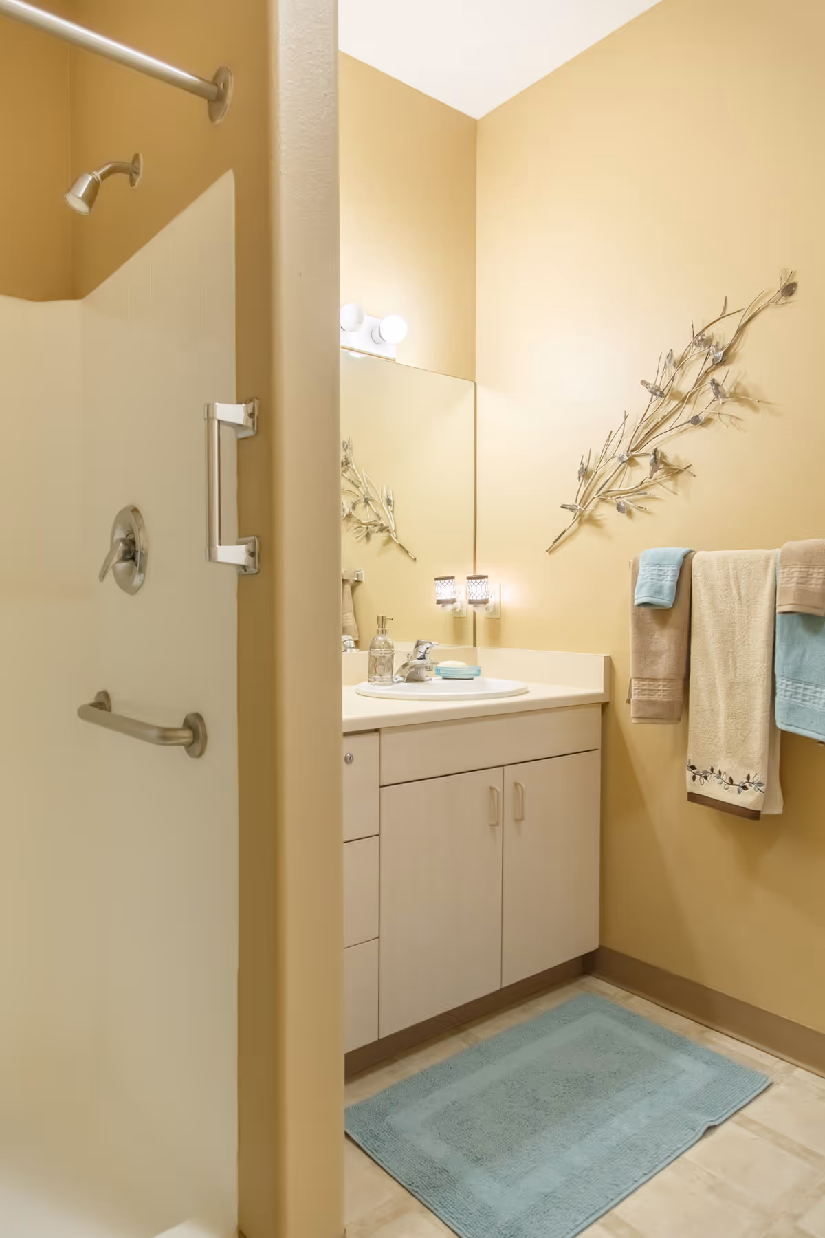A bathroom with beige walls featuring a shower area with a silver showerhead and grab bar, a vanity with a sink, soap dispenser, and a bar of soap. Above the vanity is a large mirror with two light bulbs. On the wall next to the vanity, there is a decorative branch and a towel rack holding four towels in beige, cream, and light blue colors. A light blue bath mat is on the floor.