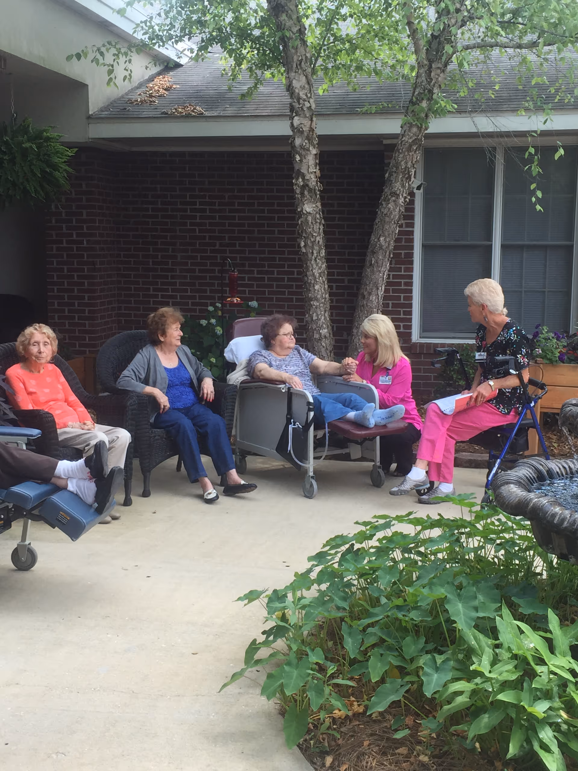 A group of elderly women sitting outdoors in a courtyard area of a nursing center. They are seated in chairs and a wheelchair, engaged in conversation. There are trees, plants, and a brick building with windows in the background.