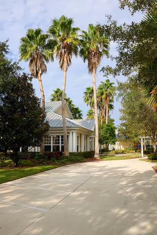 A paved driveway leading to a single-story building with a metal roof, surrounded by tall palm trees and other greenery under a partly cloudy sky.
