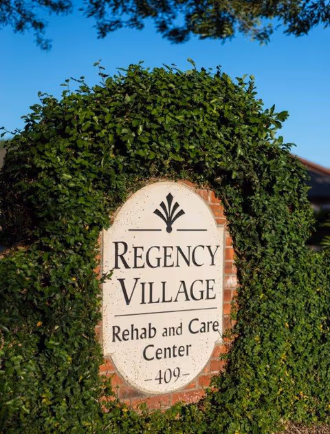 Entrance sign reading "Regency Village Rehab and Care Center" set in a brick inset surrounded by green foliage.