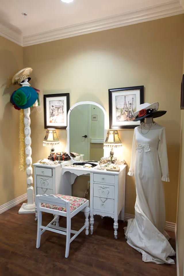 A vintage white vanity with mirror and lamps, a floral bench, a hat stand and a mannequin wearing a white dress arranged against a beige wall.