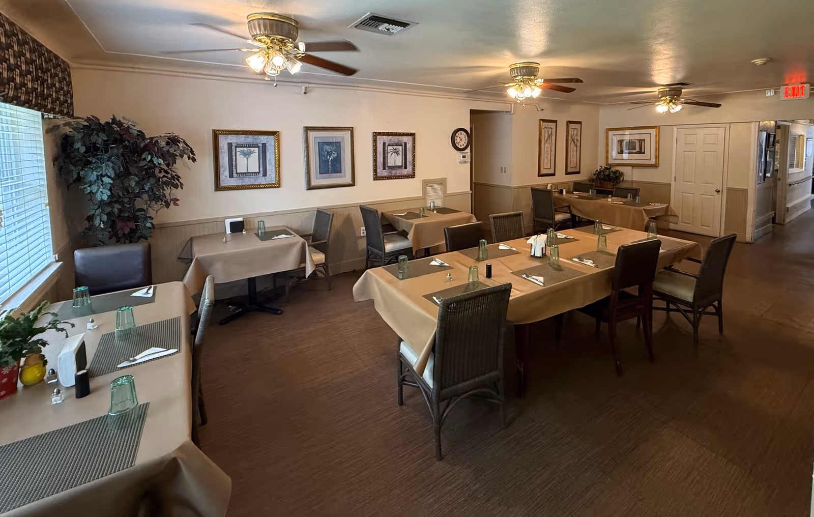Dining room with several tables covered in beige tablecloths, each set with green glasses, napkins, and utensils. The room has ceiling fans with lights, framed artwork on the walls, and large windows with blinds letting in natural light. There are chairs around the tables and some plants in the corners.
