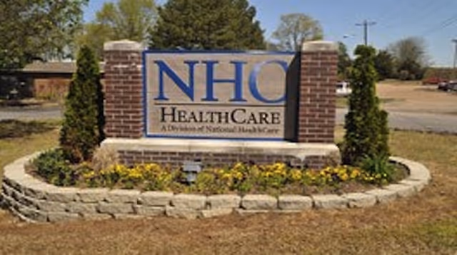 Outdoor view of a brick and stone sign for NHC HealthCare, surrounded by small shrubs and flowers, with a grassy area and trees in the background.
