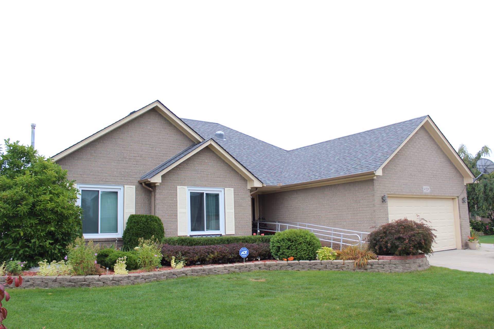 Single-story brick ranch-style house with attached garage, front lawn, shrubs, and a wheelchair ramp leading to the entrance.