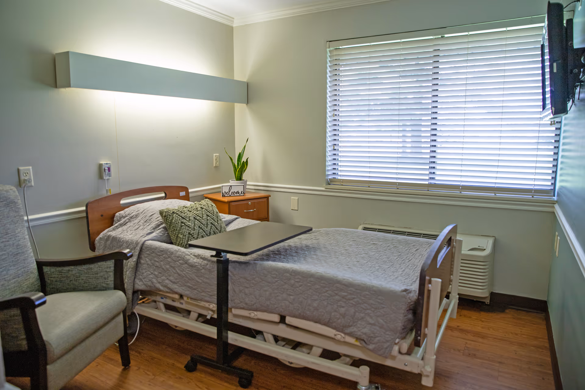 A nursing facility bedroom with a hospital bed covered in a gray quilt, a green patterned pillow, a small overbed table, a wooden nightstand with a potted plant and a welcome sign, a cushioned armchair, a large window with closed blinds, and a wall-mounted television.