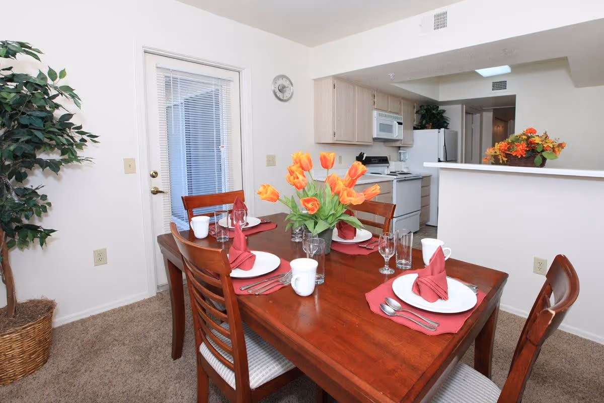 A dining area with a wooden table set for four people, featuring white plates, red napkins, glasses, and mugs. A vase with orange tulips is placed in the center of the table. The dining area is adjacent to a kitchen with light wood cabinets, a white stove, microwave, and refrigerator. A potted plant is in the corner near a door with blinds, and a basket with flowers is on the kitchen counter.