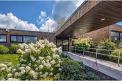 Front entrance of a brick nursing facility with a covered drop-off, an accessibility ramp, American flags, and white flowering shrubs under a partly cloudy sky.