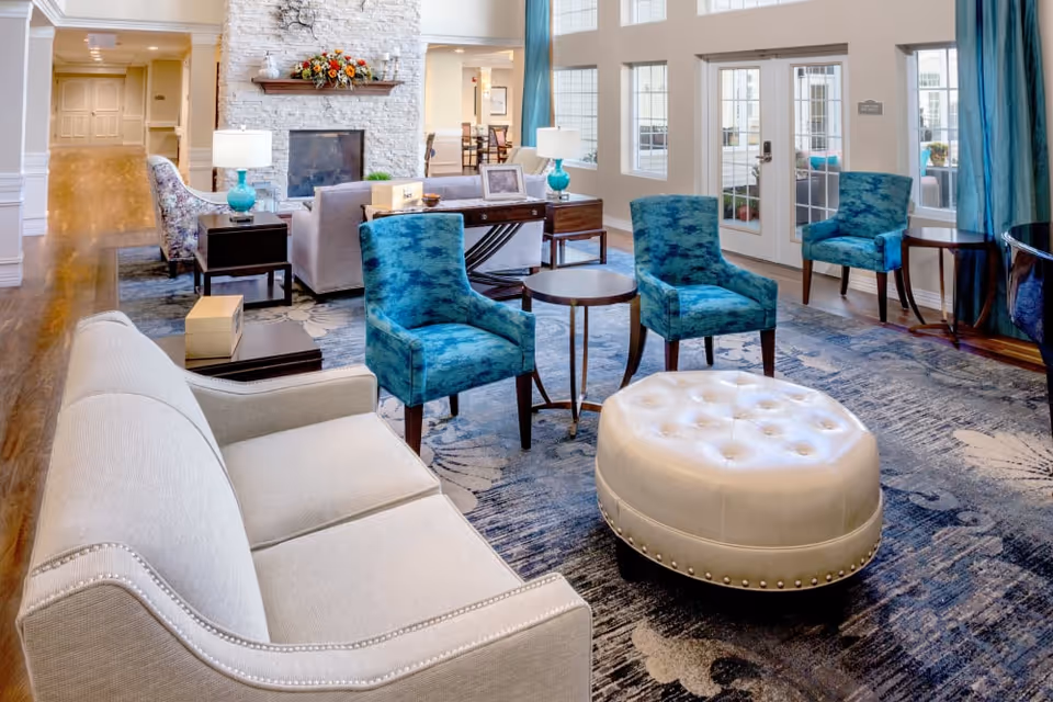 A bright and spacious living room area in an assisted living facility featuring a beige sofa, three blue upholstered chairs, a round white tufted ottoman, and several small wooden tables. The room has large windows and glass doors letting in natural light, a stone fireplace with a floral arrangement on the mantel, and hardwood floors with a patterned area rug.