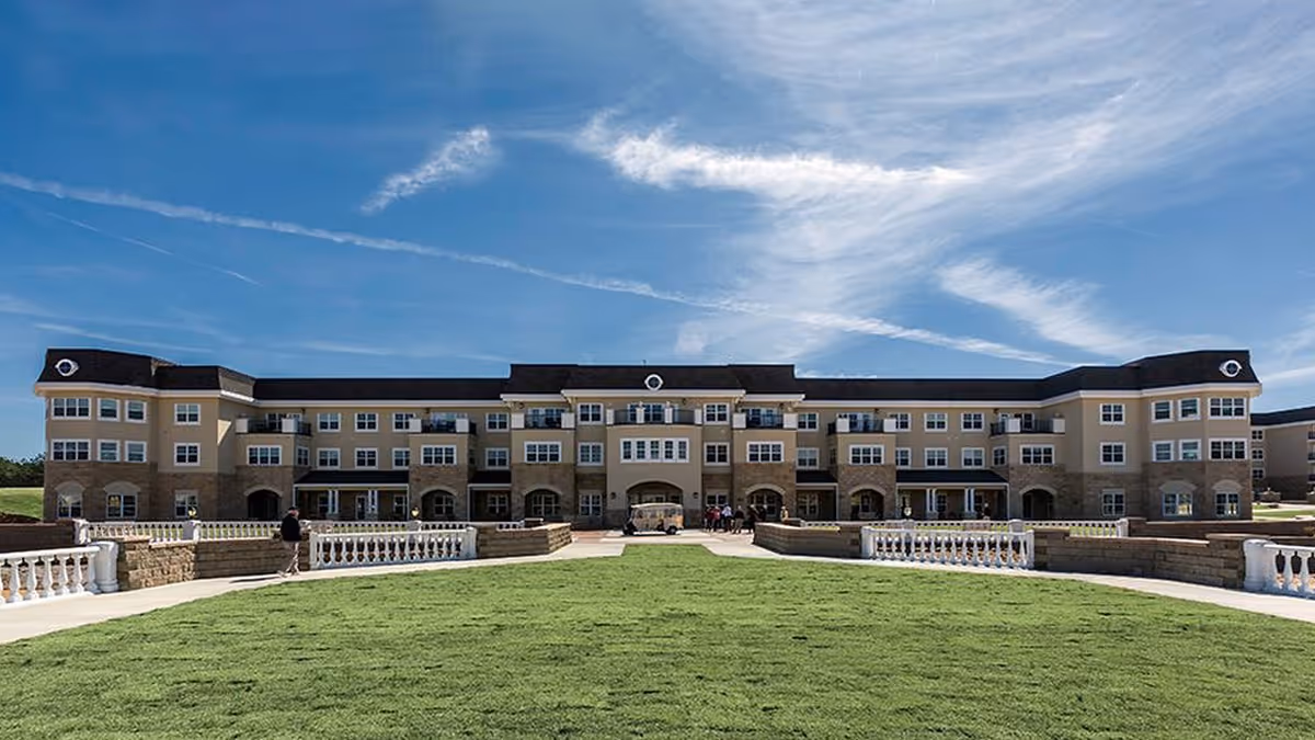 Wide exterior view of a large, three-story retirement community building with beige and stone facade under a partly cloudy blue sky. In front of the building is a well-maintained green lawn bordered by white railings and stone walls, with a few people walking near the entrance.