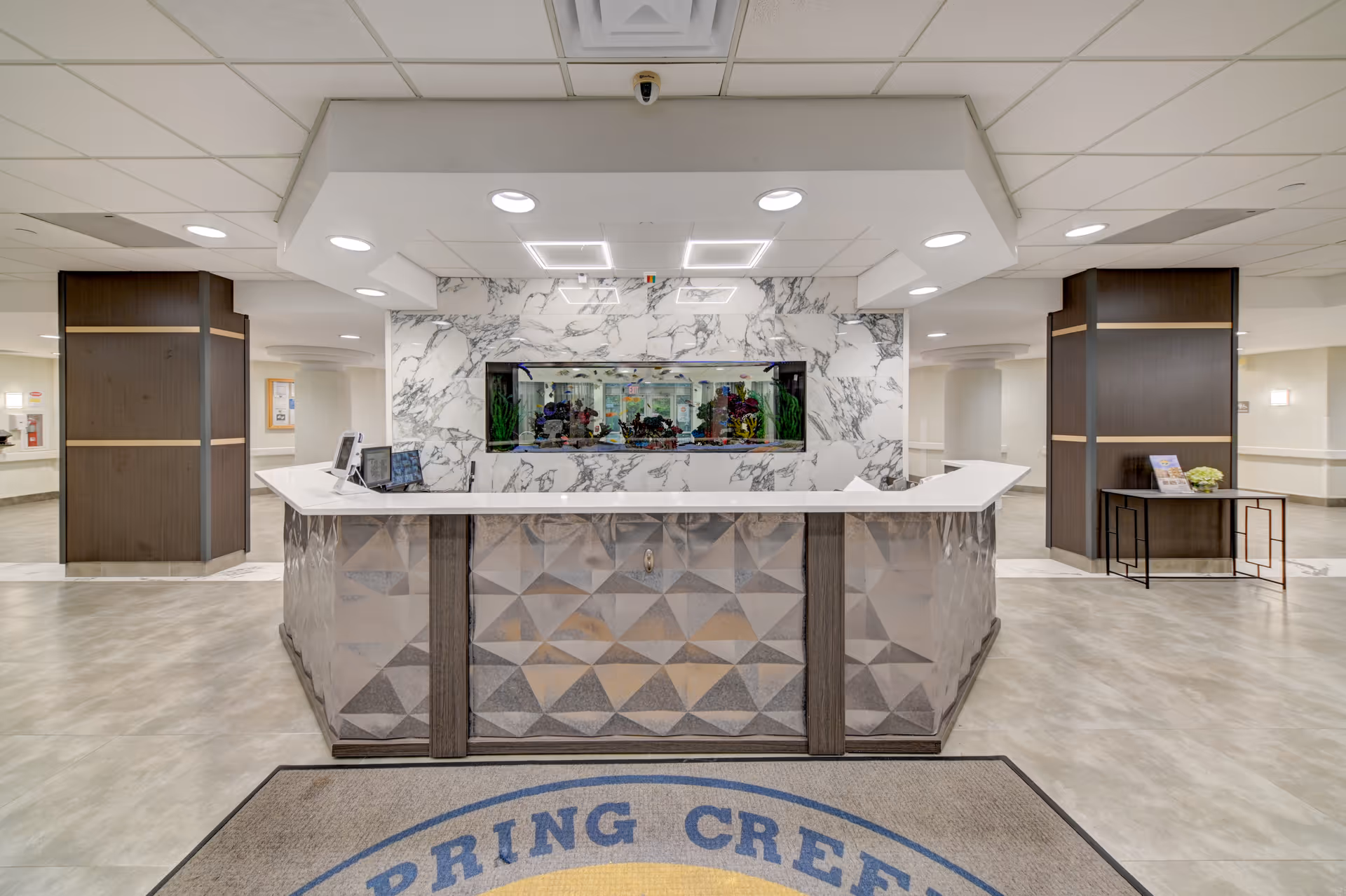 Reception desk area inside a facility with a modern design featuring a geometric patterned front desk, a large built-in aquarium behind the desk, marble wall accents, and overhead lighting. The floor has a mat with the partial text 'SPRING CREEK'.