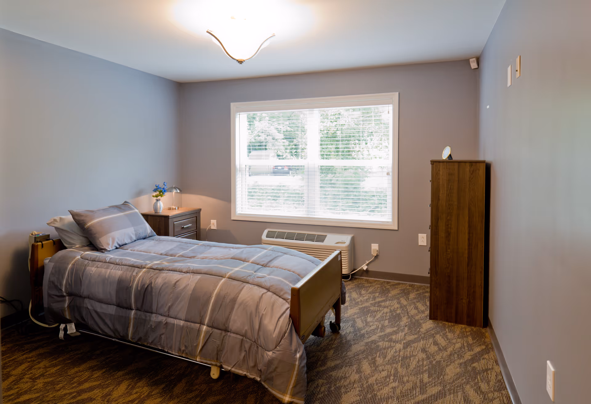 A simple bedroom in an assisted living facility with a single bed covered in a gray comforter, a wooden nightstand with a small lamp and a vase with blue flowers, a window with white blinds letting in natural light, and a wooden dresser against the wall.