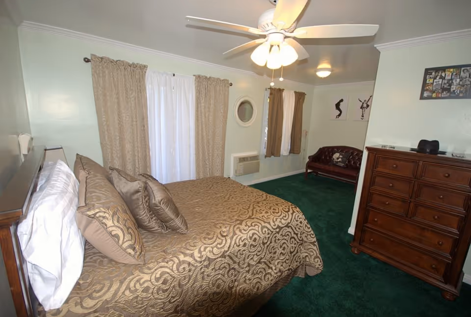 A bedroom with a large bed covered in a gold and brown patterned bedspread and matching pillows. The room has green carpet, light-colored walls, two windows with beige curtains, a ceiling fan with lights, a wooden dresser, and a small brown leather couch. There are framed pictures on the walls.