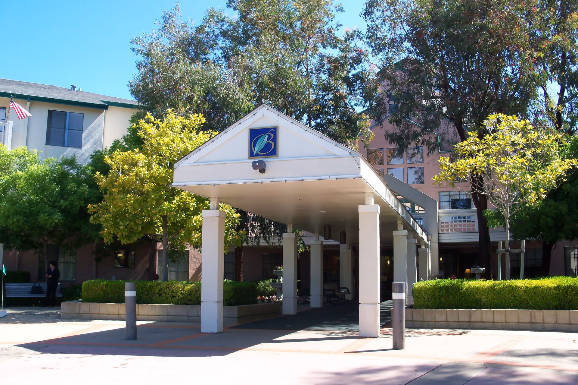 Entrance to Baywood Court facility with a covered drop-off area supported by white pillars, surrounded by green trees and bushes under a clear blue sky.