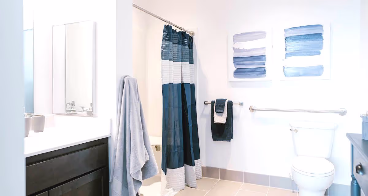 Bright accessible bathroom with a blue-striped shower curtain, vanity and mirror, towels, and a toilet with a grab bar.