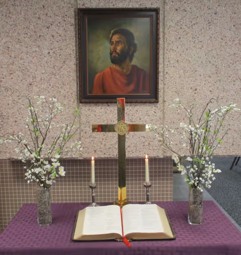 An altar table with an open Bible, a brass cross, two lit candles and vases of white flowers beneath a framed portrait of Jesus.
