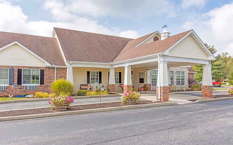 Exterior view of a single-story senior living facility building with a covered entrance supported by white columns with brick bases. The building has a combination of brick and light-colored siding, a brown shingled roof, and several windows. There are flower pots with colorful flowers near the entrance and a paved driveway in front.