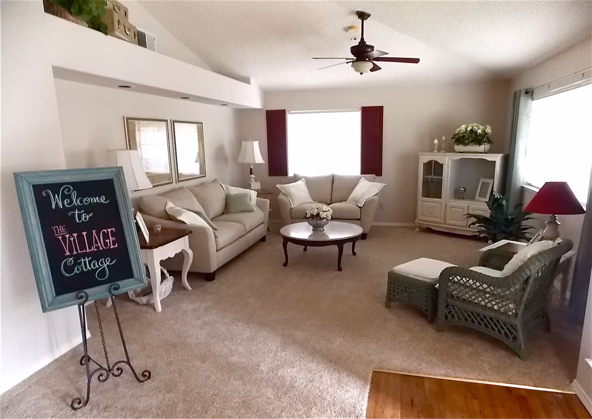 A cozy living room with beige carpet and neutral-colored furniture including a sofa, loveseat, and a wicker chair with an ottoman. A round wooden coffee table with a flower arrangement is centered in the room. There are two framed pictures on the wall above the sofa, a white side table with a lamp, and a white cabinet with decorative items. A ceiling fan is mounted on the ceiling. A chalkboard sign on a stand reads 'Welcome to The Village Cottage.'