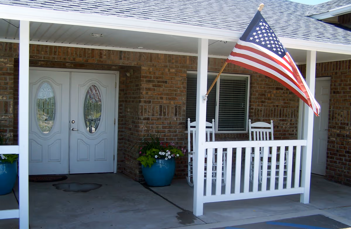 Covered front porch and entrance with white double doors, railing and rocking chairs, potted plants, and an American flag.
