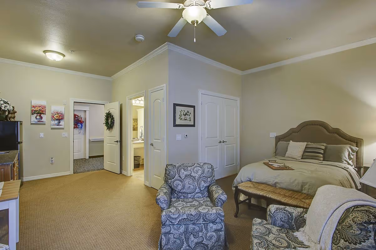A cozy bedroom in a senior living facility featuring a bed with pillows and a tray, two patterned armchairs, a ceiling fan, and an open door leading to a bathroom. The room has beige walls and carpet, with floral artwork on the wall and a small kitchenette area visible on the left.