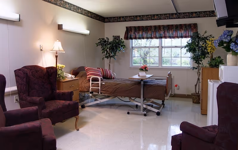 A senior living facility room with a hospital-style bed covered in brown bedding and striped pillows, two burgundy upholstered armchairs, a wooden nightstand with a lamp and a phone, a small table on wheels at the foot of the bed, potted plants near a window with floral valance curtains, and a small white refrigerator and microwave on the right side.