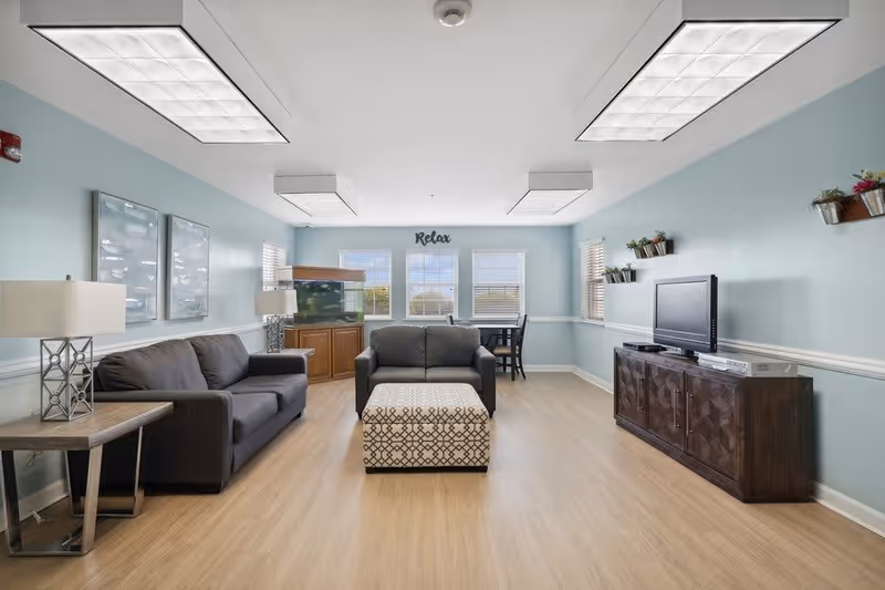 A bright and spacious living room with light blue walls and wood flooring. The room features a dark gray sofa and loveseat, a patterned ottoman in the center, a wooden TV cabinet with a flat-screen television, and a fish tank in the corner. There are two table lamps on side tables next to the sofa, wall-mounted planters with greenery, and three windows letting in natural light. The word 'Relax' is displayed above the windows.