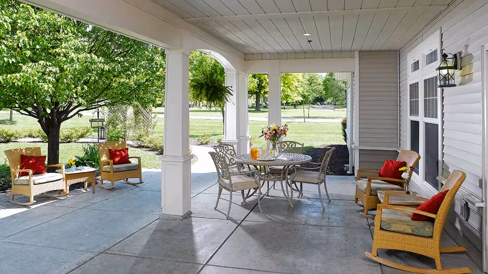Covered outdoor patio area with wicker rocking chairs and a round metal table with chairs. The patio overlooks a green lawn with trees and shrubs, and there are red cushions on the chairs and a vase with flowers on the table.