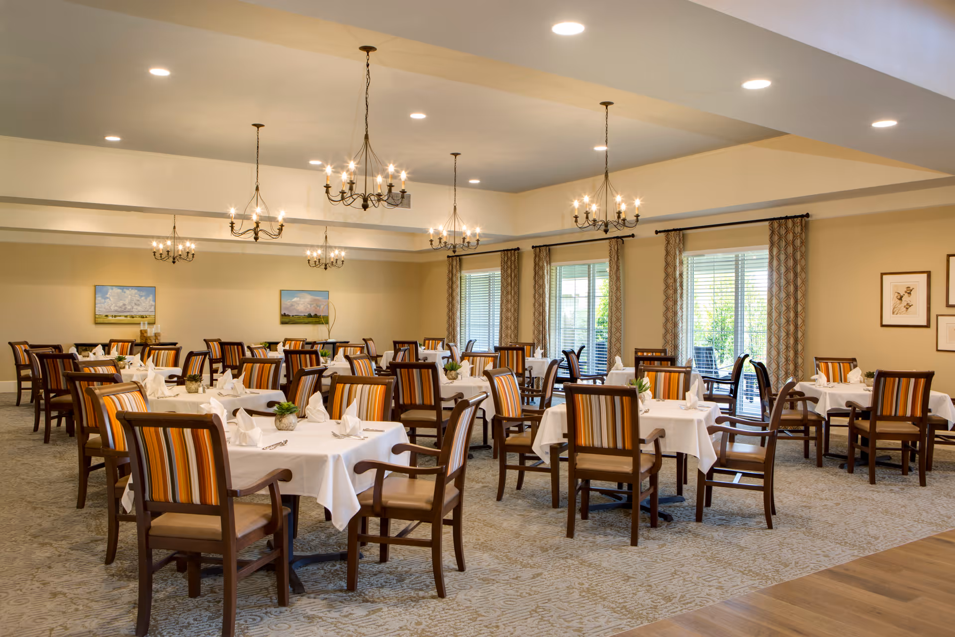 A spacious dining room with multiple tables covered in white tablecloths, each set with napkins and small potted plants. The chairs have wooden frames with striped upholstery in warm tones. The room is well-lit with chandeliers and recessed ceiling lights. Large windows with patterned curtains let in natural light, and framed artwork decorates the walls.