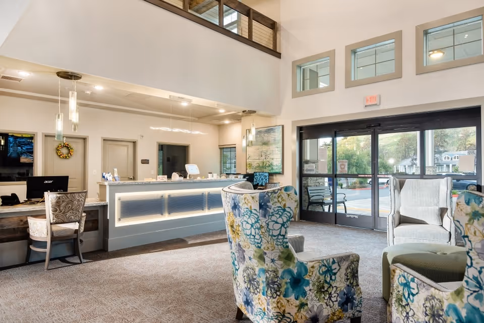 A bright and welcoming reception area with a front desk, computer, and chairs. The space features high ceilings, pendant lights, floral patterned armchairs, and large glass doors leading outside with a view of greenery and houses.