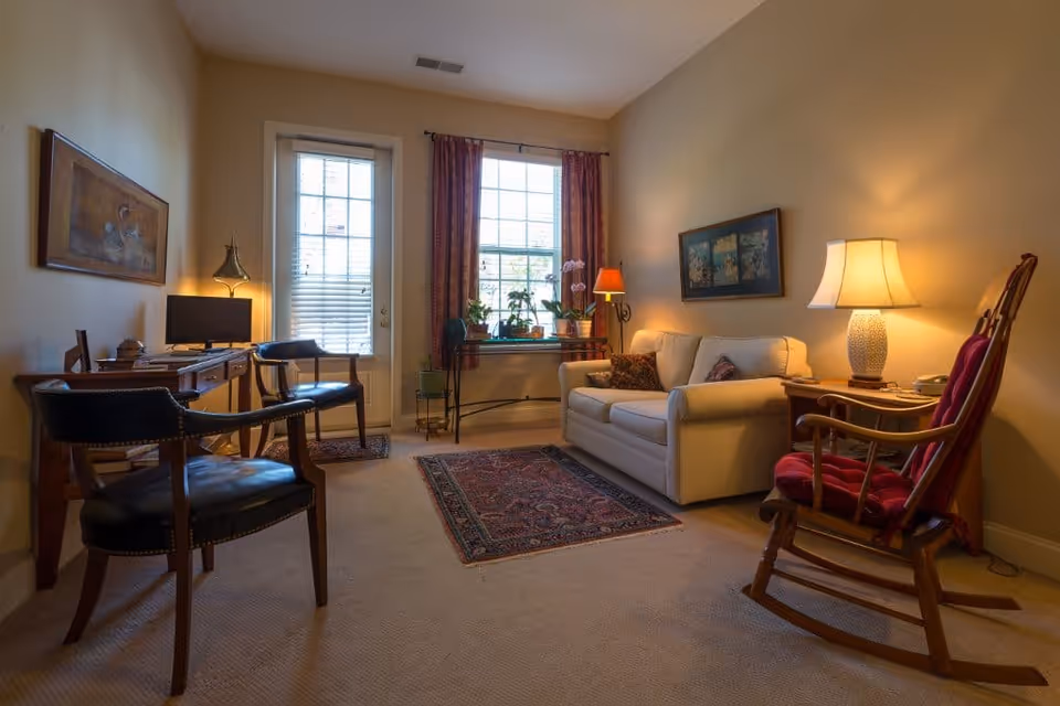 Cozy living room with a sofa, rocking chair, desk, area rug and a window with potted plants and lamps.