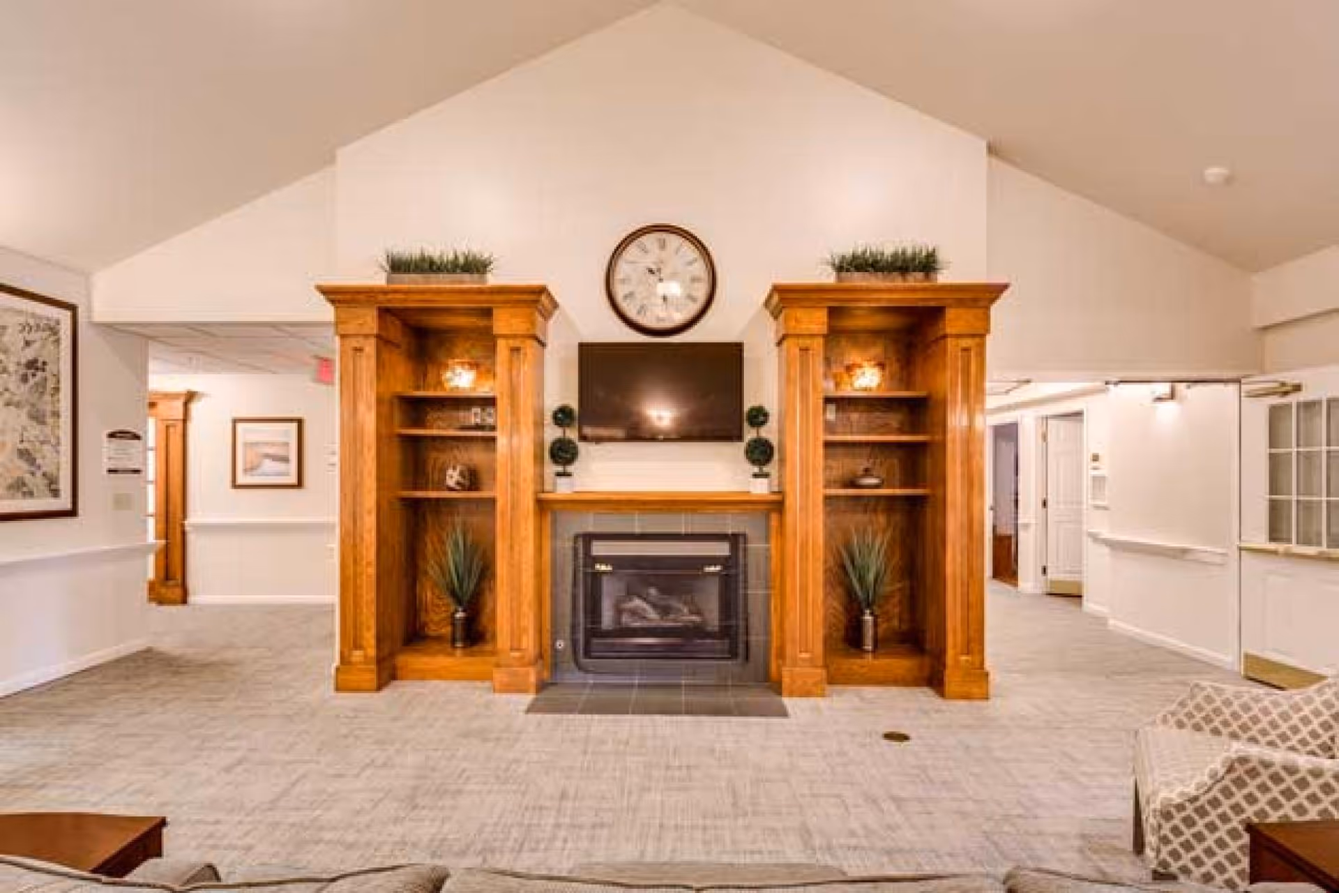 A cozy living room area with a central fireplace flanked by two wooden built-in shelves decorated with plants and small ornaments. Above the fireplace is a mounted flat-screen TV and a round wall clock. The room has light-colored walls and carpeted flooring, with a patterned armchair visible on the right side.