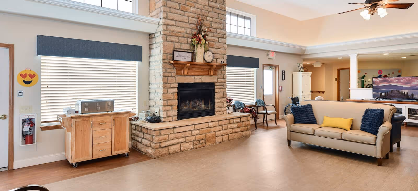 A spacious living room area in a senior living facility featuring a large stone fireplace with a wooden mantle decorated with a clock and flowers. There are two windows with blinds and blue valances on either side of the fireplace. A beige sofa with blue and yellow cushions faces a large flat-screen TV mounted on a white stand. The room has light-colored walls and flooring, with a fire extinguisher mounted near a door on the left side and a small wooden cabinet with a microwave on top. Chairs and a wheelchair are visible in the background near an exit door.
