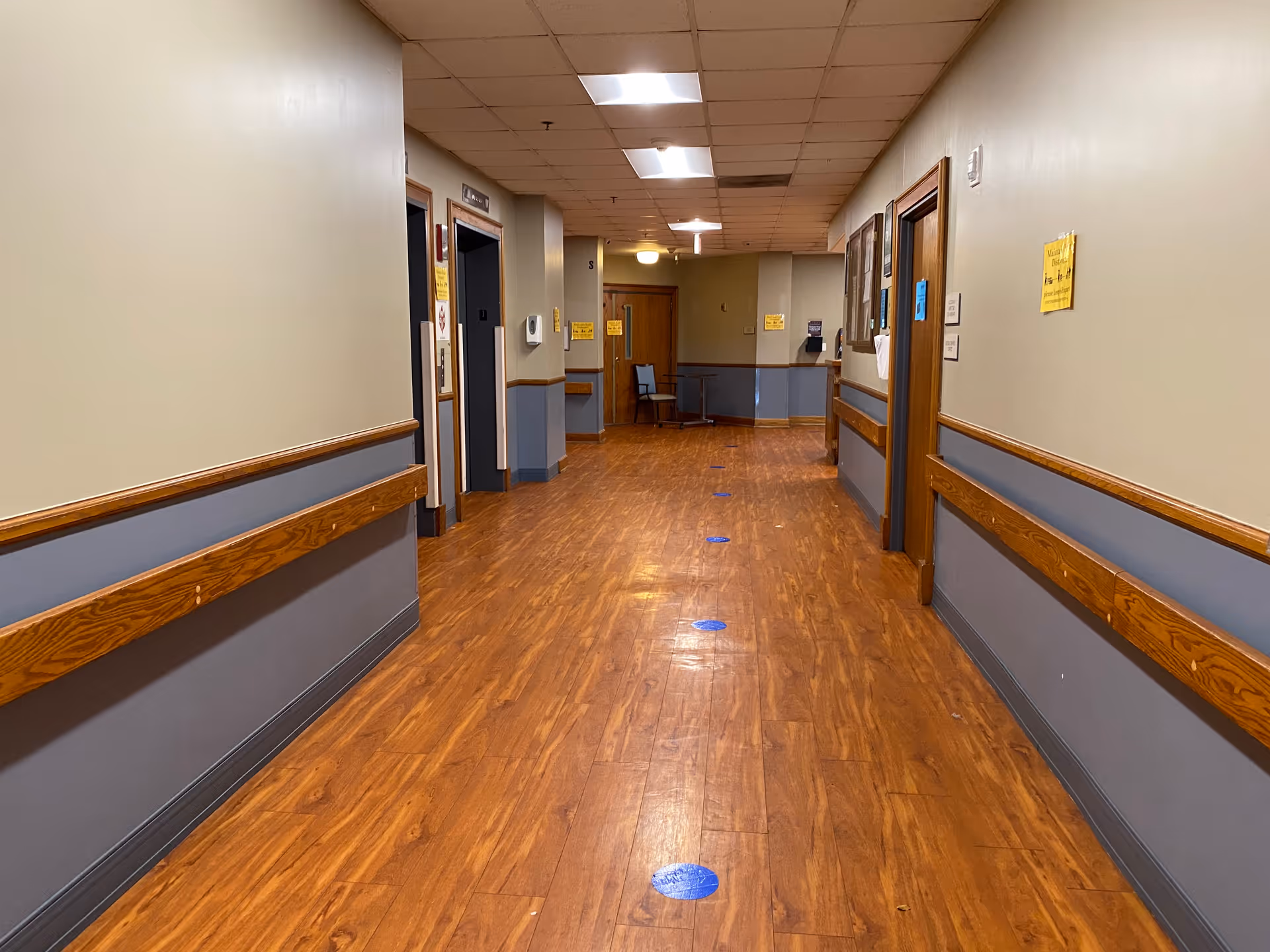 A long indoor hallway with wooden flooring and beige walls with blue lower panels. The hallway has handrails on both sides, several doors, and ceiling lights. Blue circular floor markers are spaced along the center of the hallway floor. A chair and small table are visible at the far end.