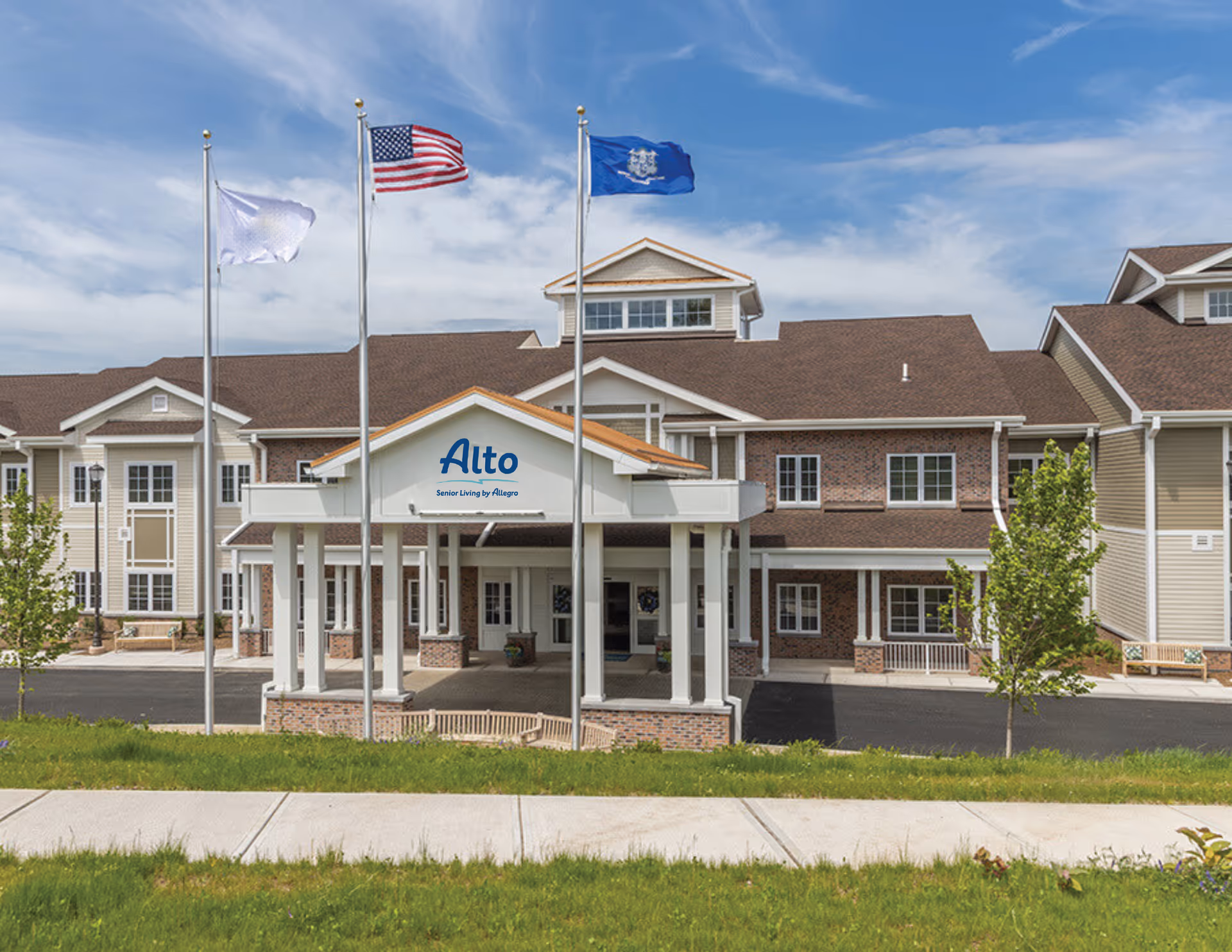 Exterior front view of Alto Evergreen Walk senior living facility with a covered entrance, three flagpoles displaying flags including the American flag, and a well-maintained lawn and sidewalk in the foreground under a partly cloudy sky.