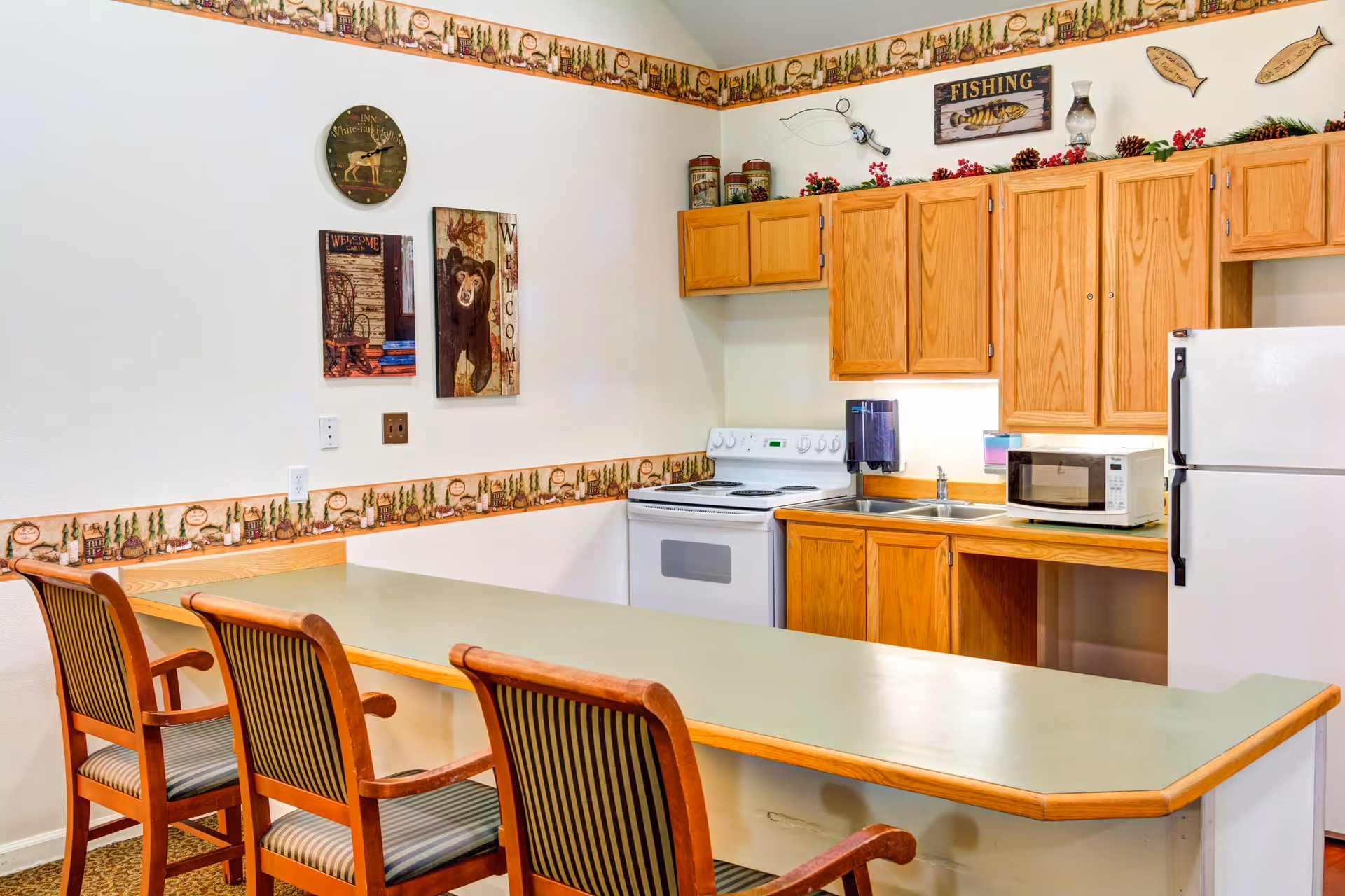 A kitchen area with wooden cabinets, a white stove, a white refrigerator, a microwave, and a double sink. There is a long counter with three wooden chairs with striped cushions. The walls are decorated with a nature-themed border and various rustic signs including a bear and fishing theme.