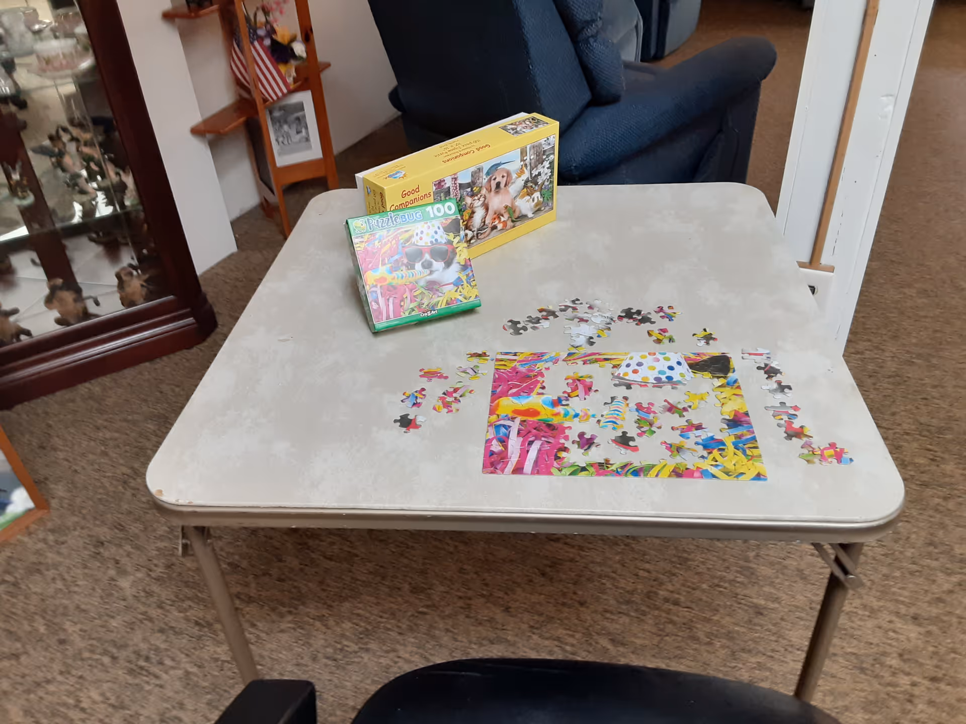 A beige folding table with a partially completed colorful puzzle and two puzzle boxes on top. The puzzle features a festive scene with bright colors and a party hat. In the background, there is a blue armchair, a wooden shelf with an American flag and framed photos, and a glass display cabinet with figurines.